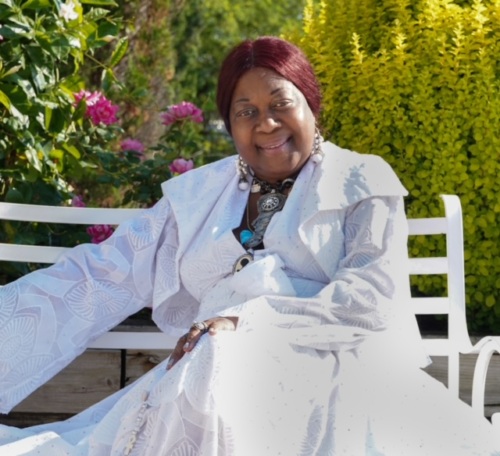 A woman in a white dress sits on a bench in a garden, surrounded by greenery and pink flowers.