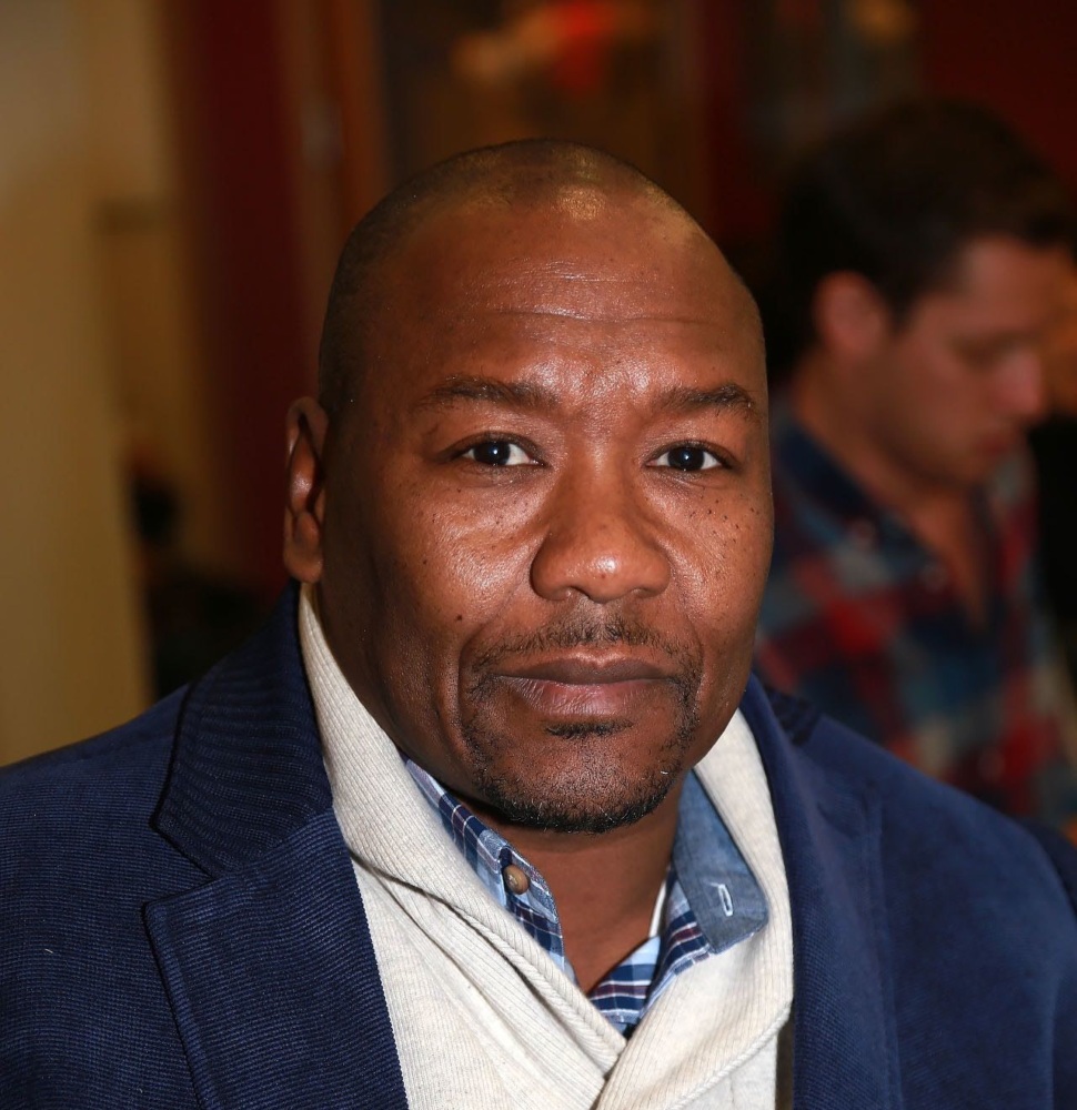 A person in a blue jacket and white scarf is looking at the camera indoors, with a blurred person in the background.