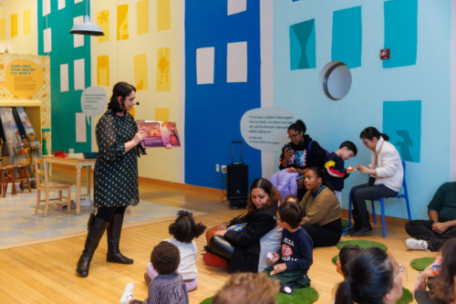 A woman reads a book to a group of children seated on the floor and adults seated on chairs in an indoor, colorful room with geometric wall designs.