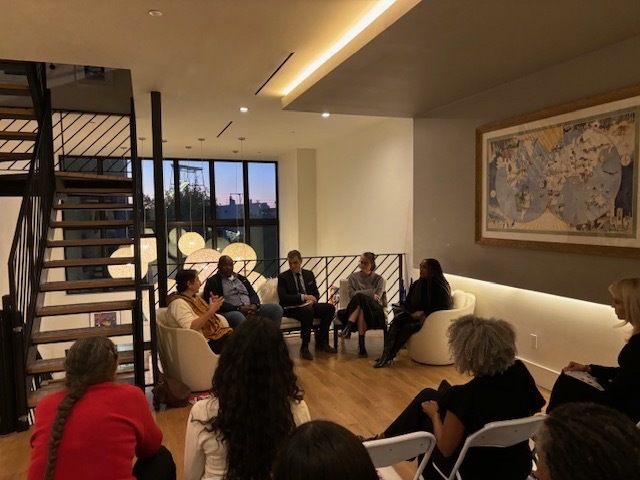 A group of people seated in a modern room are engaged in a discussion. A staircase and a large window are in the background.