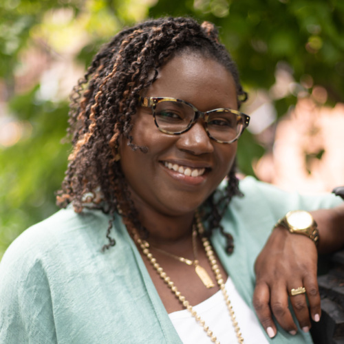 A woman wearing glasses and a green cardigan smiles while standing outdoors. She has curly hair and is wearing a large necklace.