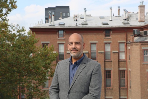 A man in a gray suit stands outdoors with a brick building and trees in the background.