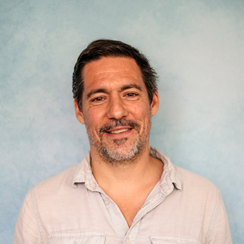 A man with short dark hair and a gray beard smiles against a light blue background, wearing a light-colored shirt.