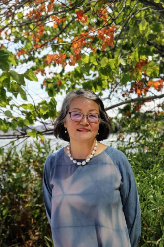 A woman in a blue dress stands outdoors with green and orange leaves in the background. She is smiling and wearing glasses and a pearl necklace.