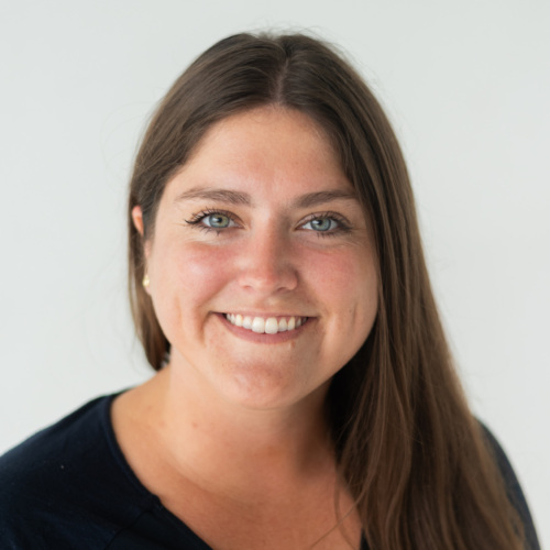 A woman with long brown hair, wearing a navy blue top, smiles at the camera against a plain white background.