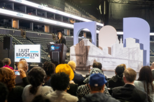 A speaker at a podium addresses an audience in an arena. A large backdrop displays a stylized Brooklyn Bridge. People in the audience are taking photos.