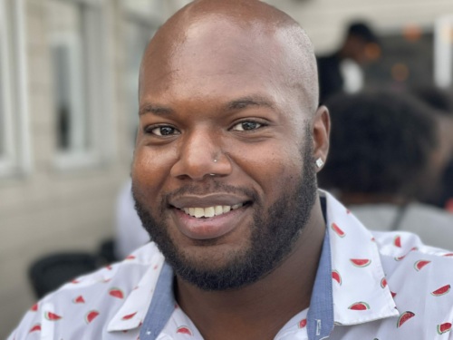 A smiling person wearing a white shirt with a watermelon pattern is outdoors, with a blurred background.