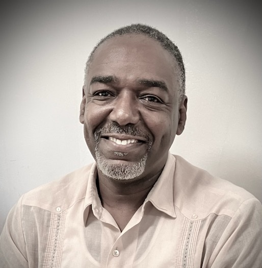 A man with short gray hair and a goatee smiles, wearing a light-colored button-up shirt, sitting against a plain background.