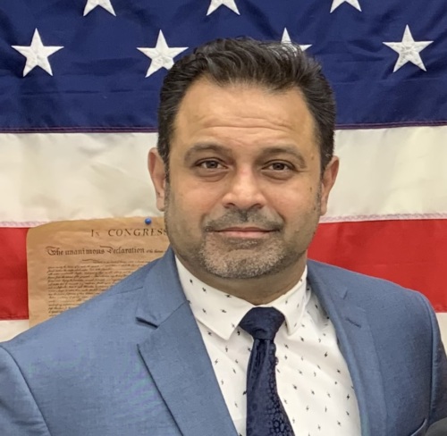 A man in a blue suit and tie stands in front of an American flag.