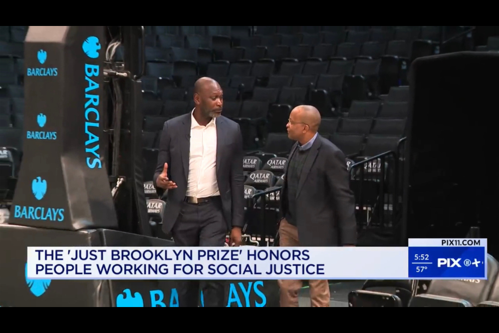 Two men in suits walk and talk on a basketball court near a news headline about the "Just Brooklyn Prize" for social justice.