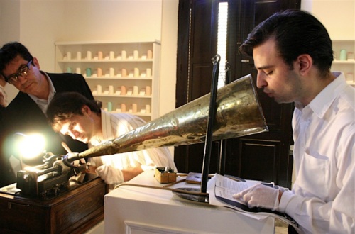 Three men engage with an antique phonograph; one speaks into a large horn, another observes, and the third adjusts the device. They are in a room with shelves of jars in the background.