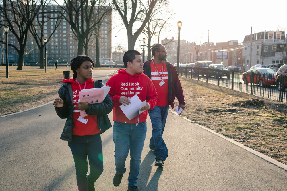 Three people wearing red "Red Hook Community Resilience" shirts walk along a park path, holding papers, with buildings and trees in the background.