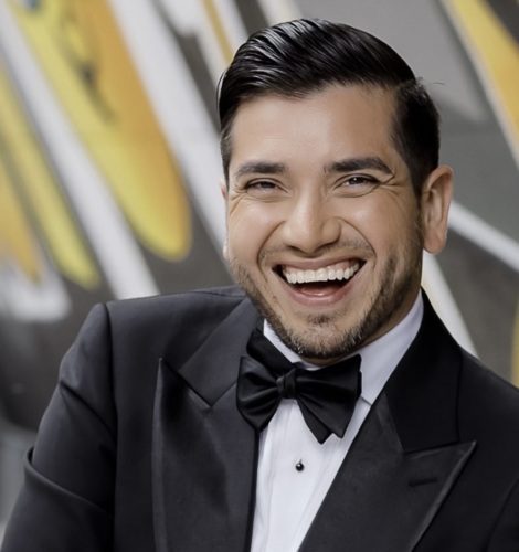 A man in a tuxedo with a bow tie smiles in front of an abstract, colorful background.