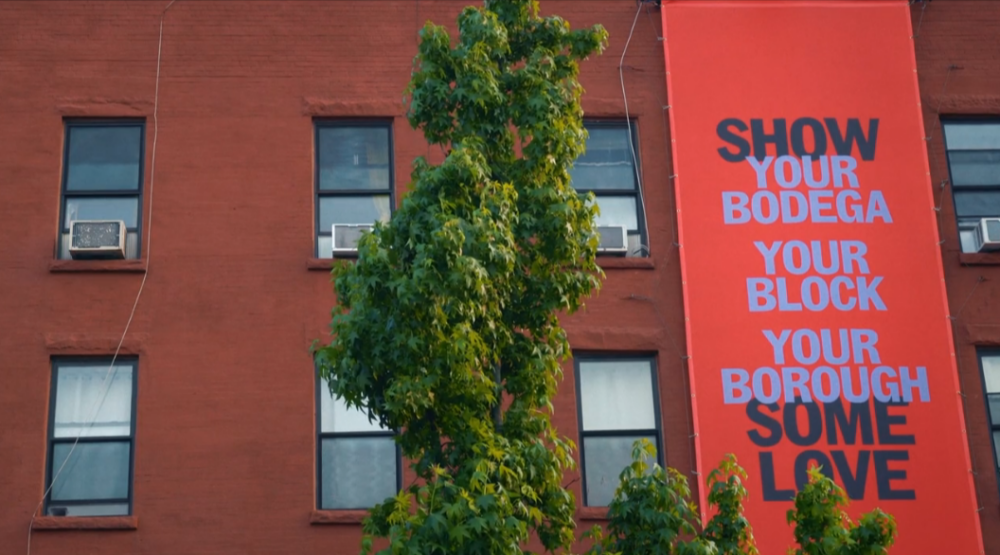 Brick building with a banner reading "SHOW YOUR BODEGA YOUR BLOCK YOUR BOROUGH SOME LOVE" next to a tree.
