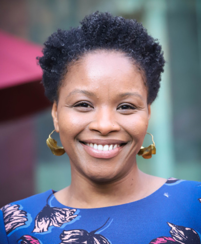 A person with short curly hair, wearing dangly earrings and a floral top, smiles broadly.