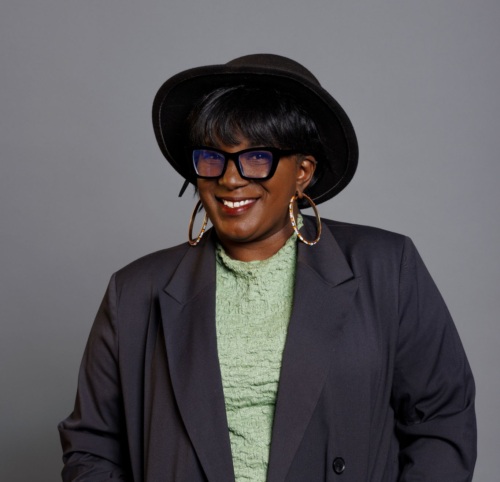 Person wearing a green dress, black overcoat, black hat, and large hoop earrings, standing against a gray background.