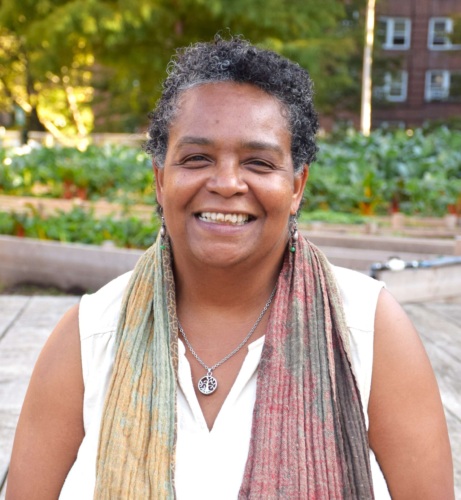 A person smiling outdoors with a colorful scarf and necklace, standing in front of a garden with trees and buildings in the background.