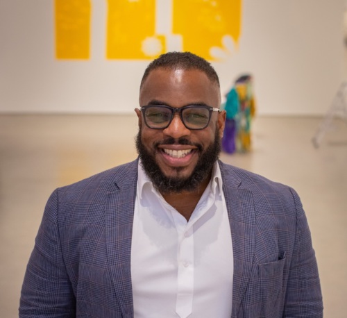 A person wearing a blue blazer, white shirt, and glasses, smiling with hands in pockets, stands in a gallery with yellow artworks in the background.