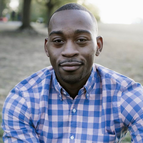 A person wearing a blue and pink checkered shirt is sitting outdoors with a slight smile, and trees are visible in the background.