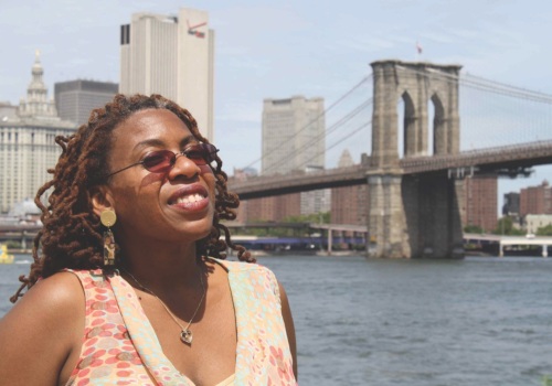 A person with sunglasses and braided hair smiles in front of the Brooklyn Bridge with city buildings in the background.