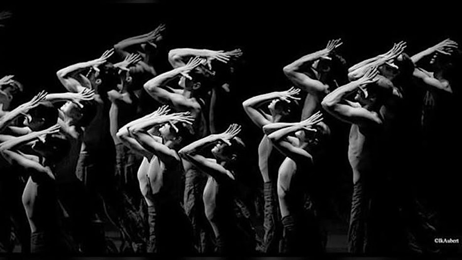 A group of dancers perform in unison, adjusting their arms above their heads, in a black and white image.