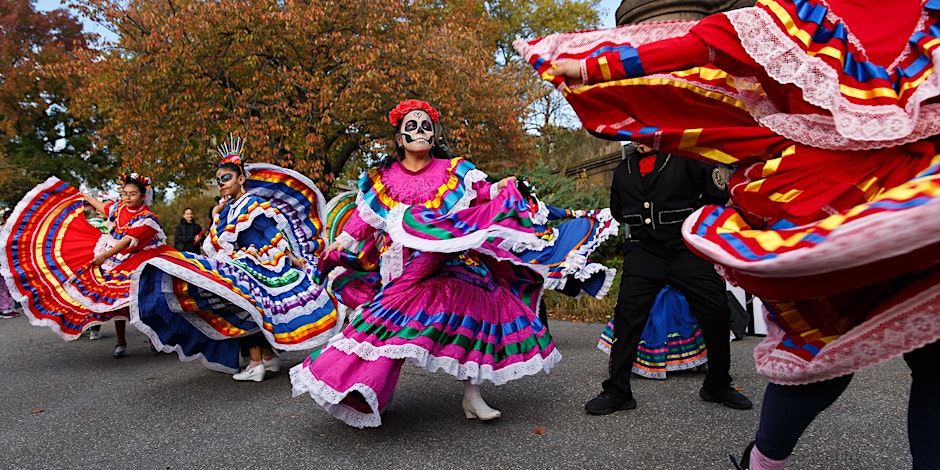 People in colorful traditional dresses dance on a street, celebrating with face paint in a festive outdoor setting.