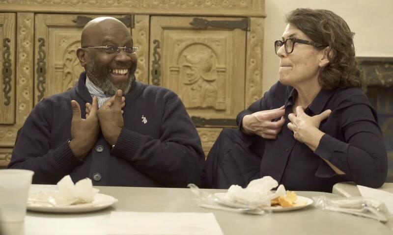 Two people sitting at a table, smiling and gesturing during a conversation. Food wrappers are visible in front of them.