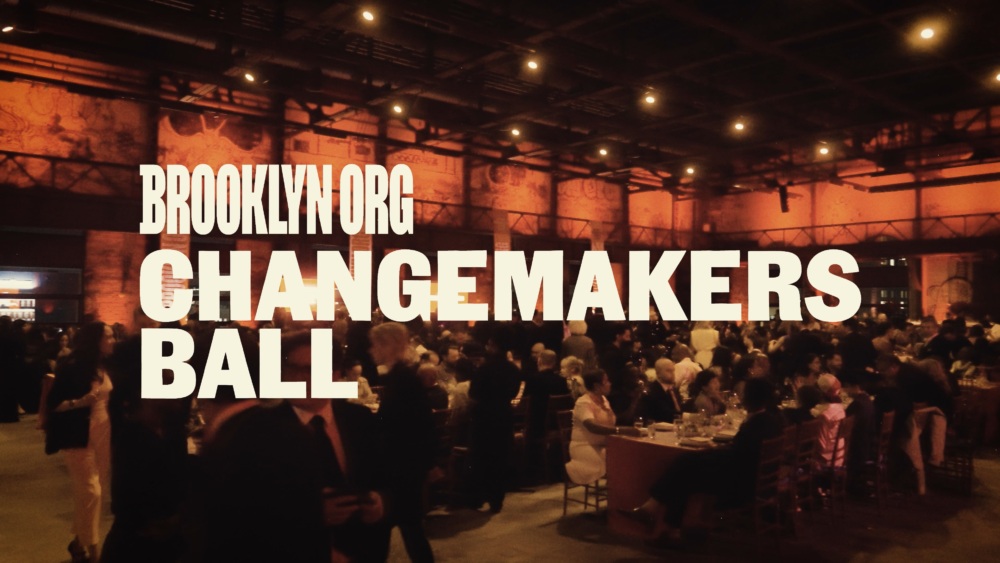 People seated and standing at a formal event with large text overlay: "Brooklyn.org Changemakers Ball." The venue is dimly lit with warm lighting.