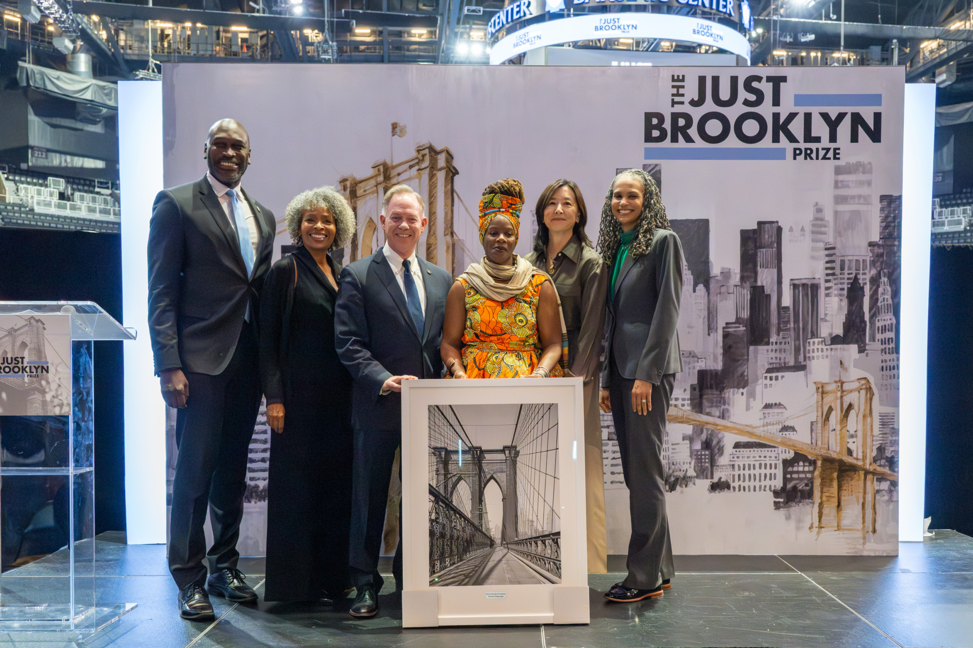 A group of seven people stands with a framed image of the Brooklyn Bridge during the Just Brooklyn Prize event.