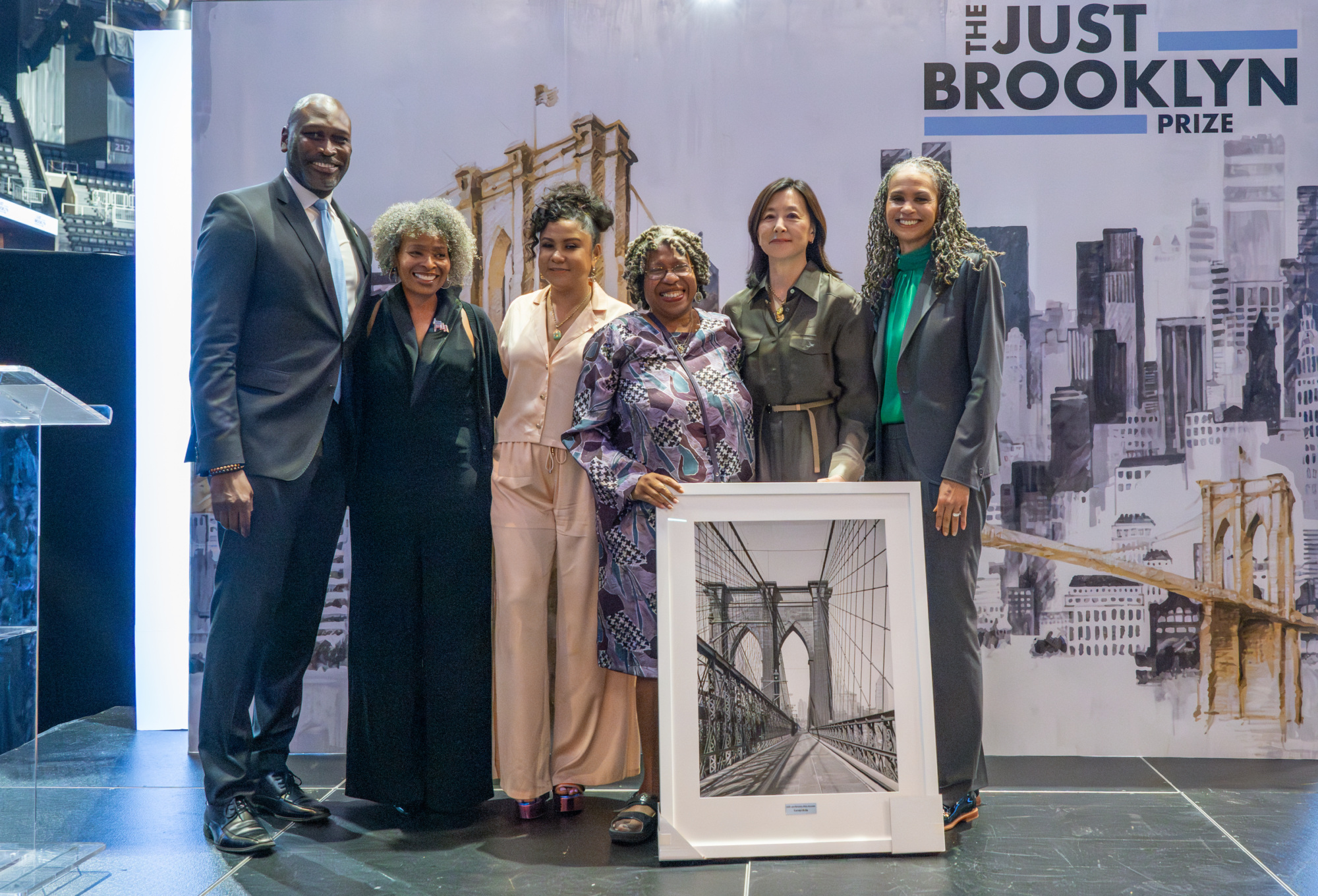 A group of six people stand together on a stage holding a framed photo of the Brooklyn Bridge. They are dressed formally, and the backdrop reads "The Just Brooklyn Prize.