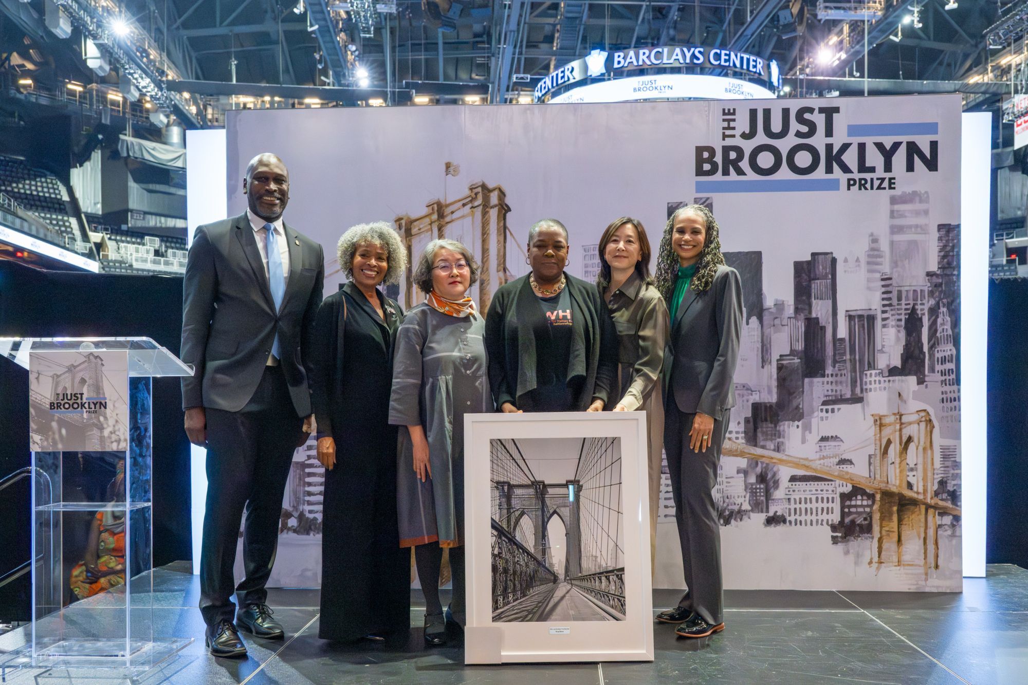 A diverse group of six people stands on a stage at the Barclays Center, holding a framed photo of the Brooklyn Bridge, with "The Just Brooklyn Prize" banner behind them.