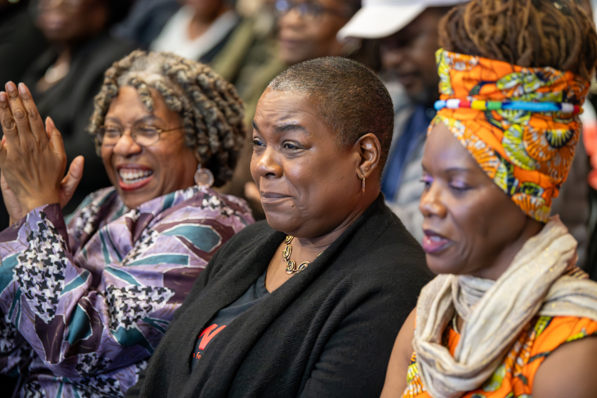 Three women sit side by side in a crowd, two smiling and one appearing focused. The setting is lively and colorful.