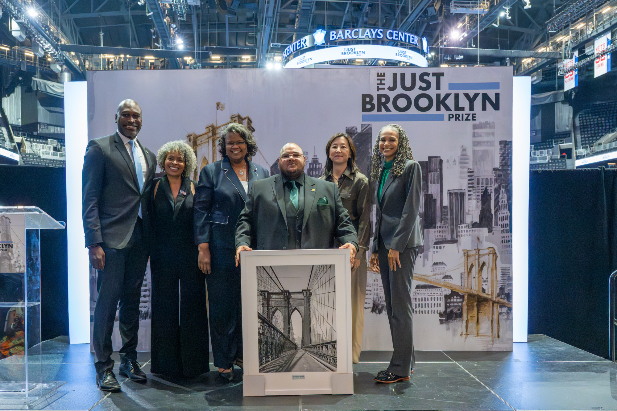 Six people pose indoors holding a framed Brooklyn Bridge artwork, with a large "Just Brooklyn Prize" backdrop behind them.