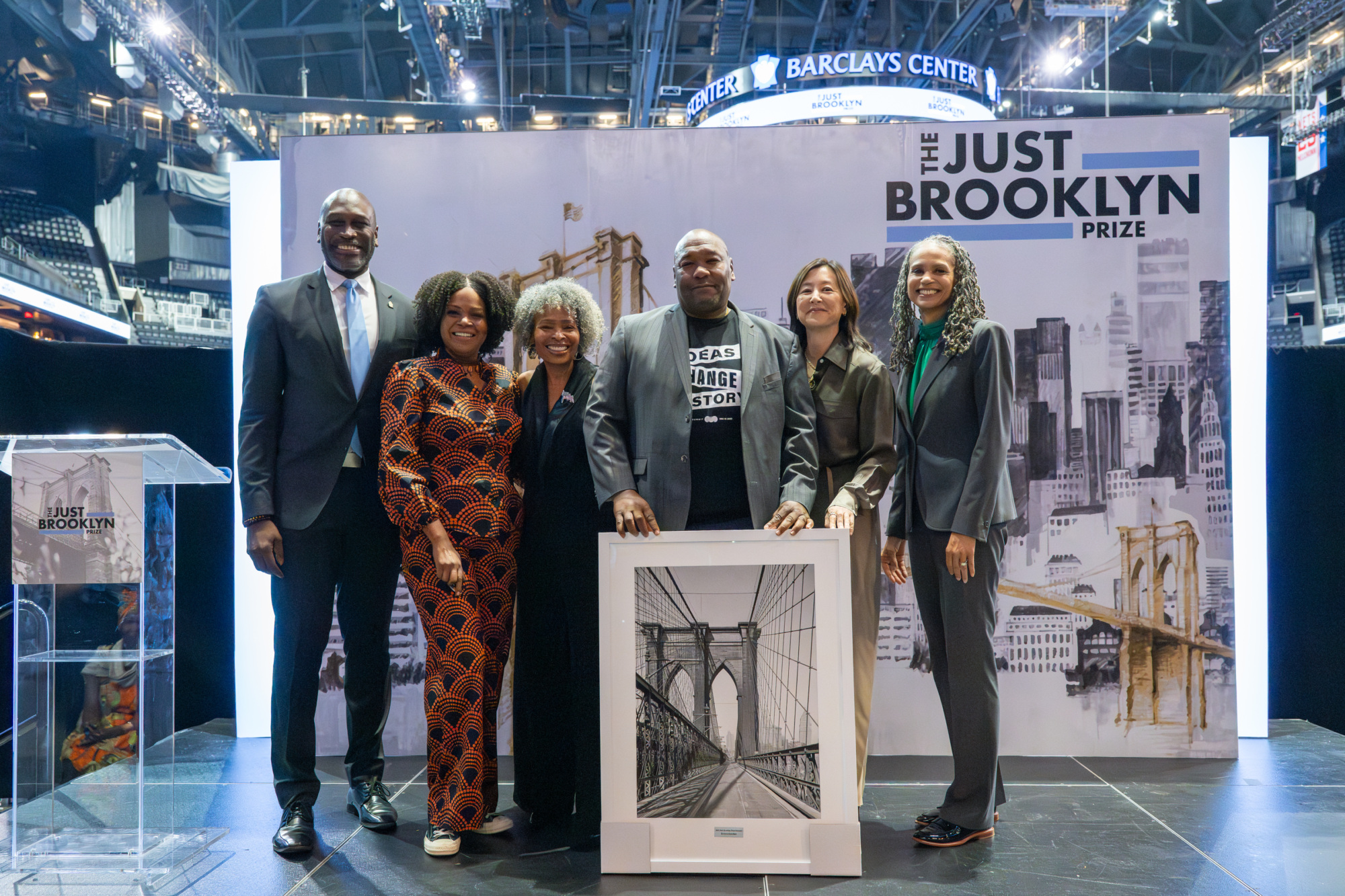 A group of people stands on stage holding a Brooklyn Bridge photo at the Just Brooklyn Prize event in Barclays Center.