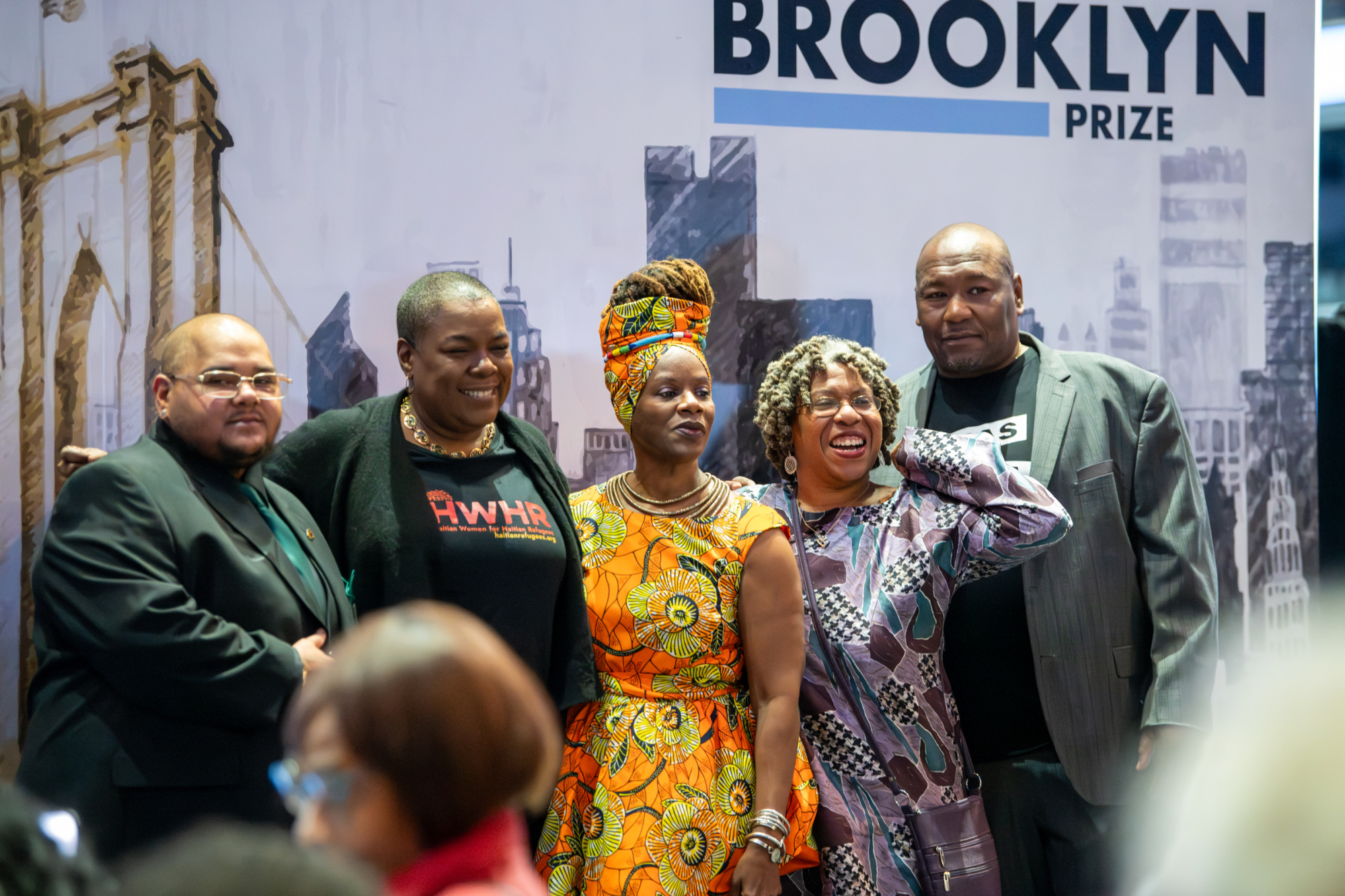 Five people stand together in front of a backdrop with "Brooklyn Prize" written on it.