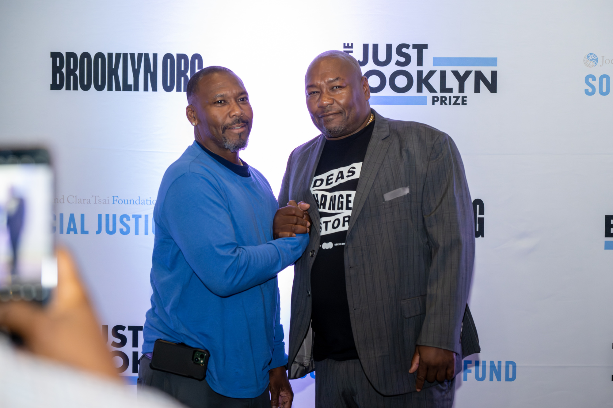 Two men are posing together in front of a backdrop displaying "Brooklyn Org" and "The Just Brooklyn Prize." One is wearing a blue shirt, the other a gray suit.