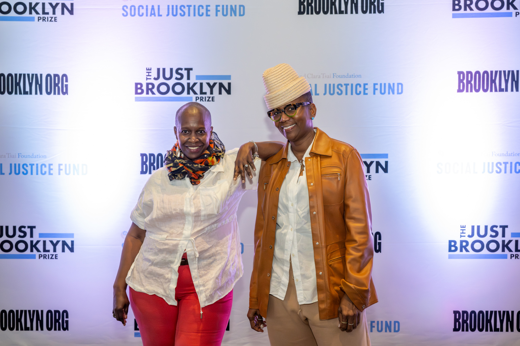 Two people smiling and posing in front of a backdrop featuring logos for The Just Brooklyn Prize and Social Justice Fund.