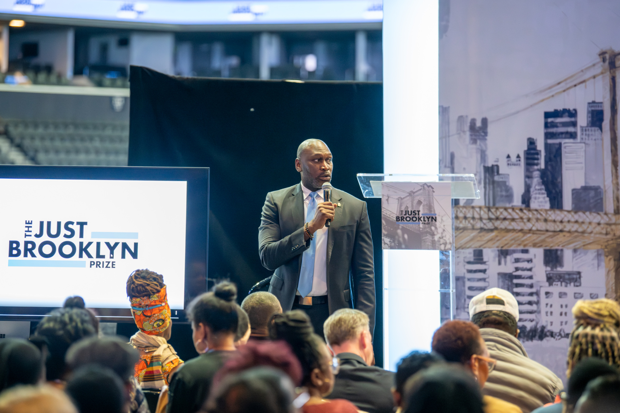 A person speaks into a microphone on stage at an event, with a "The Just Brooklyn Prize" sign on a podium. Audience members are seated in the foreground.