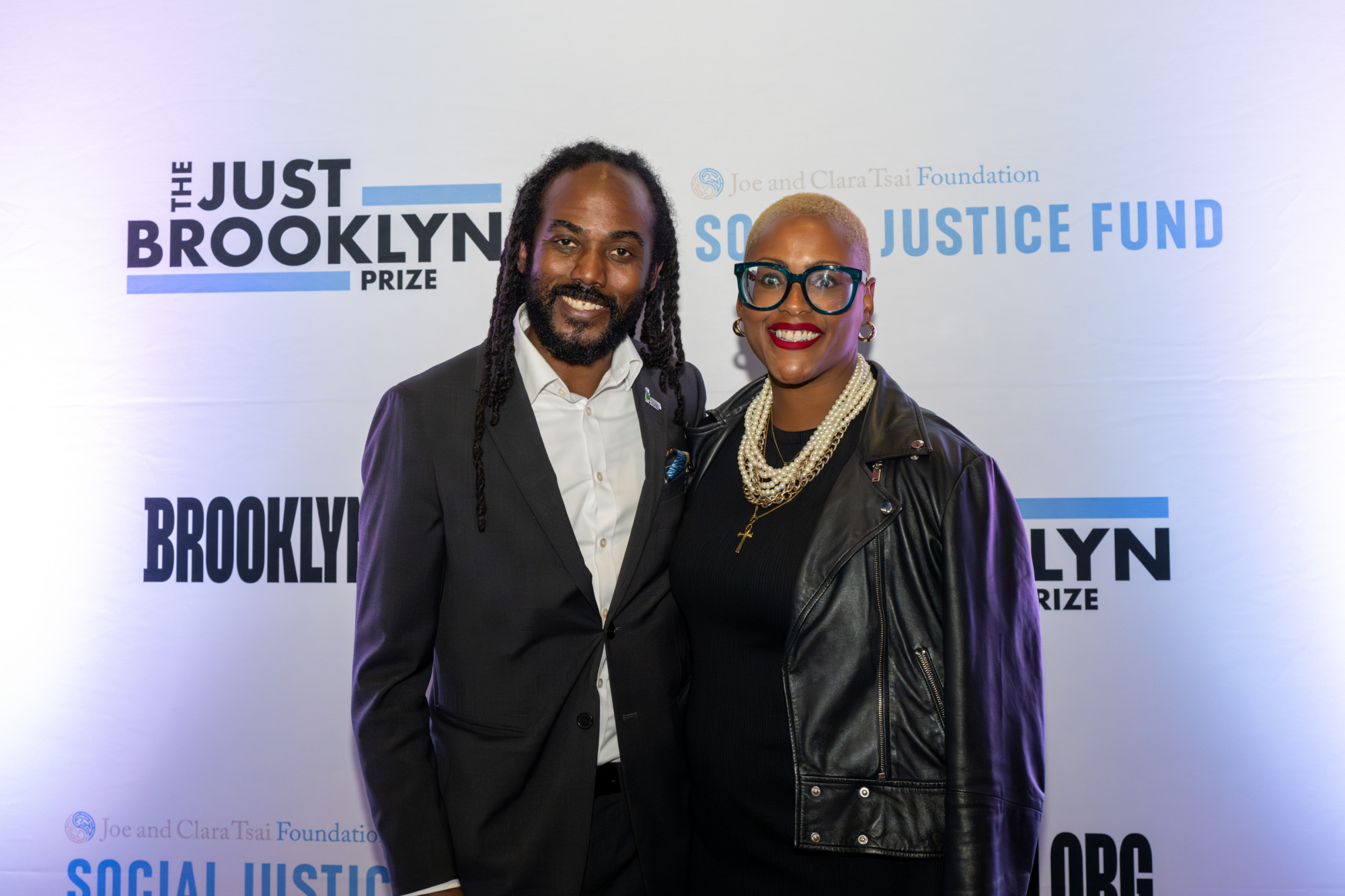 A man and woman pose together in front of a backdrop with the text "The Just Brooklyn Prize" and "Social Justice Fund.