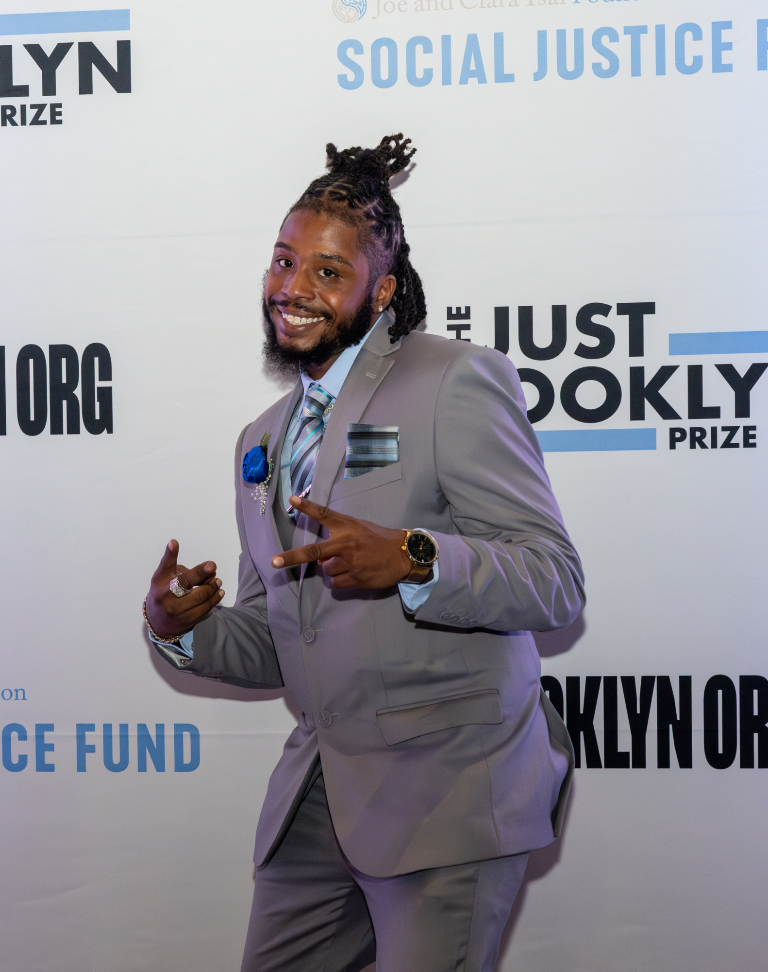 A person in a gray suit poses with peace signs on a red carpet labeled "Brooklyn Prize" and "Social Justice Fund.