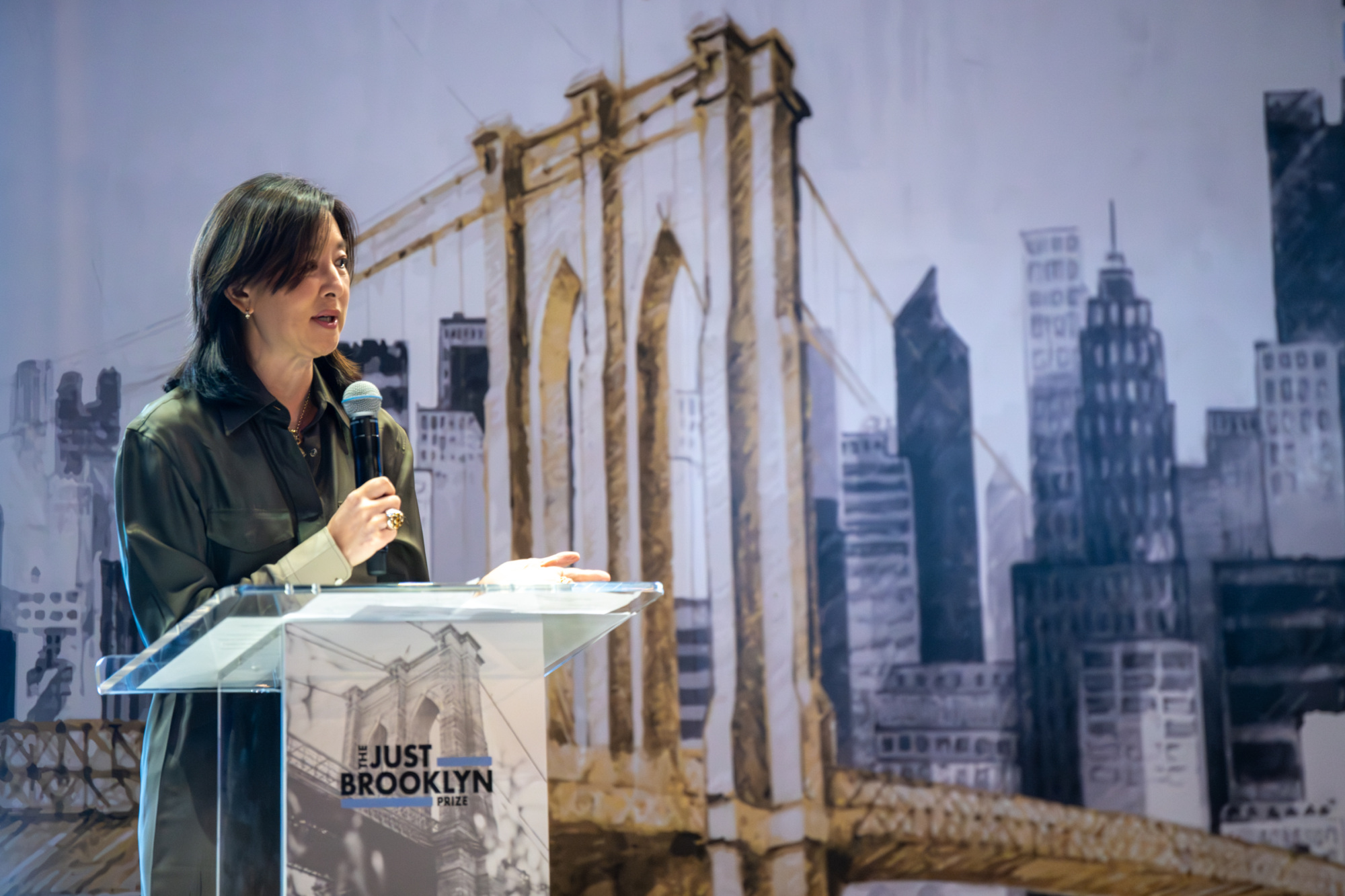 A person speaks at a podium with a microphone. A mural of the Brooklyn Bridge and cityscape is in the background.