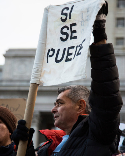 An individual holds a sign reading "SI SE PUEDE" at a public gathering, wearing a black jacket and gloves. A building is visible in the background.