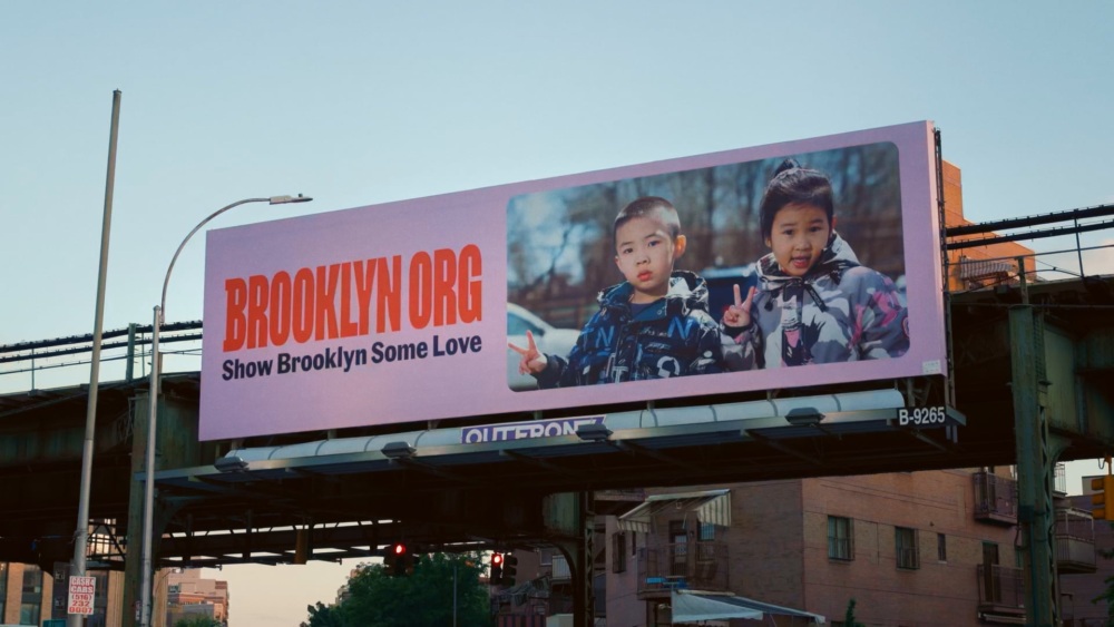 Billboard with two children posing, one boy and one girl. Text reads "BROOKLYN.ORG Show Brooklyn Some Love." Mounted on an overpass with buildings in the background.