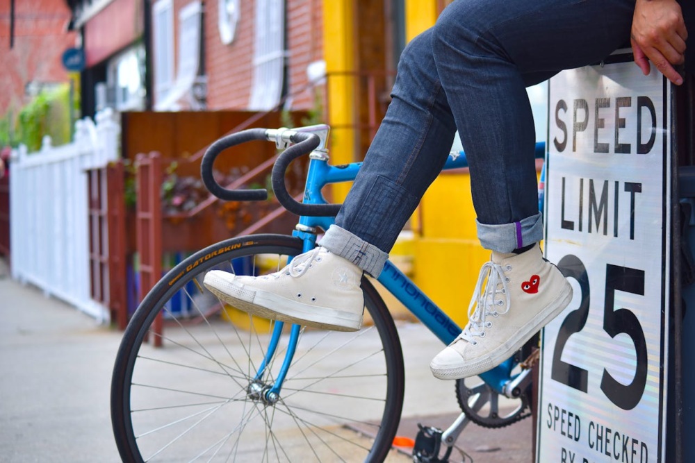 Person sitting on a speed limit sign with legs crossed, wearing white high-top sneakers with a red heart design, next to a blue bicycle on a street.