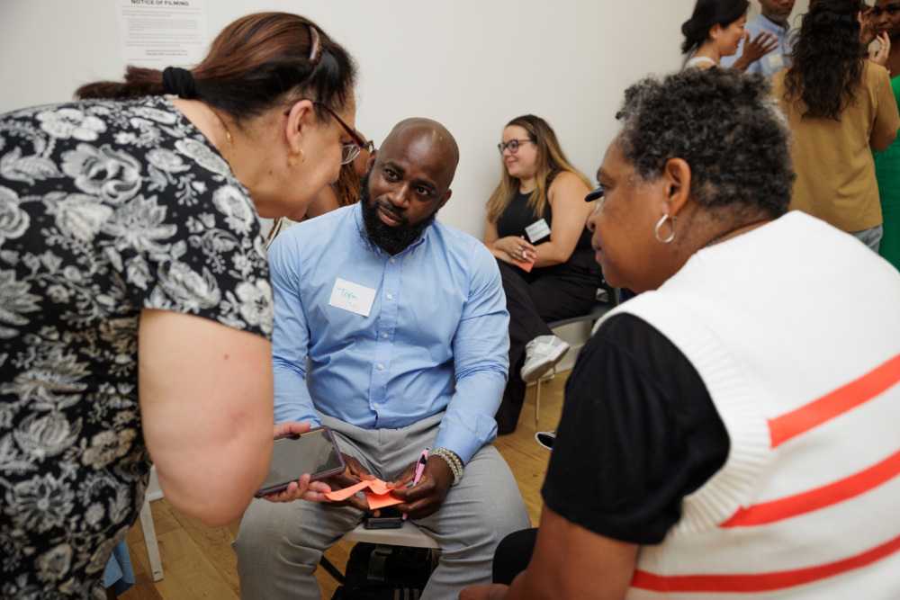 Three people are sitting and having a discussion in a room. One person is holding a phone. Other people are visible in the background.