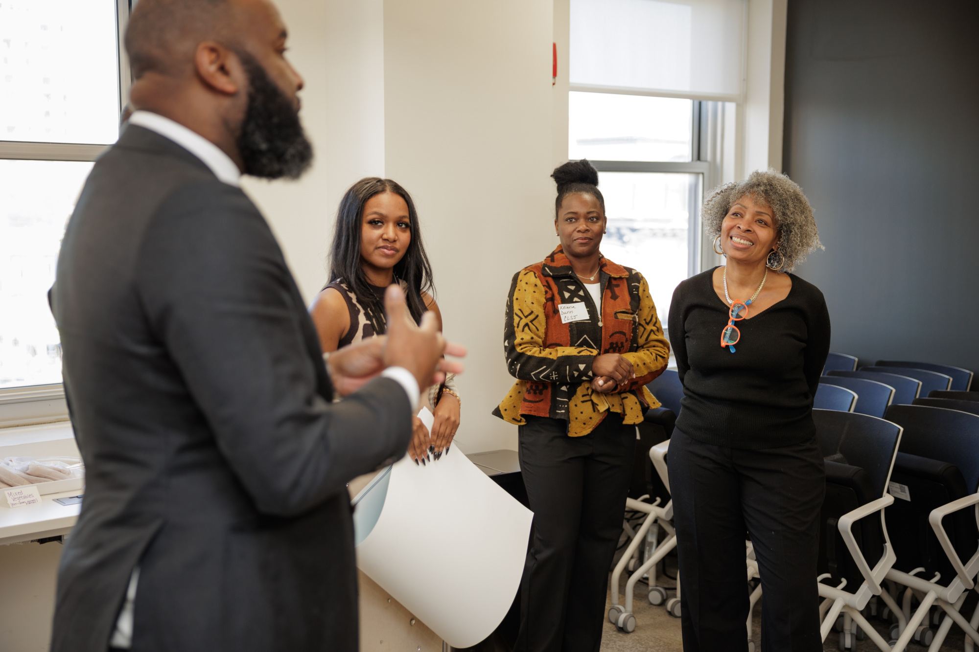 A group of four people is having a conversation in an office setting. Three women listen attentively to a man speaking. They are standing near a stack of folded chairs.