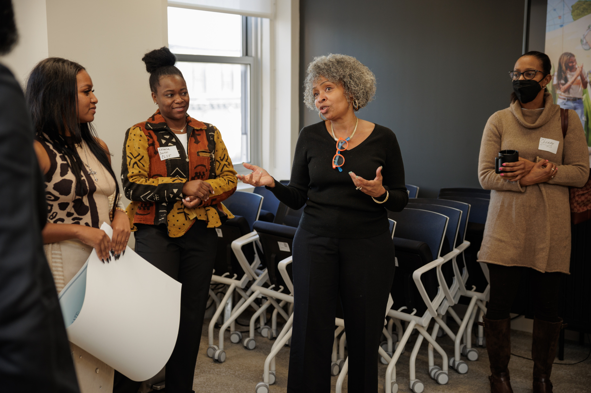 A woman speaks to a group in an office setting. Three other women listen, one holding a cup. Stacked chairs are in the background.