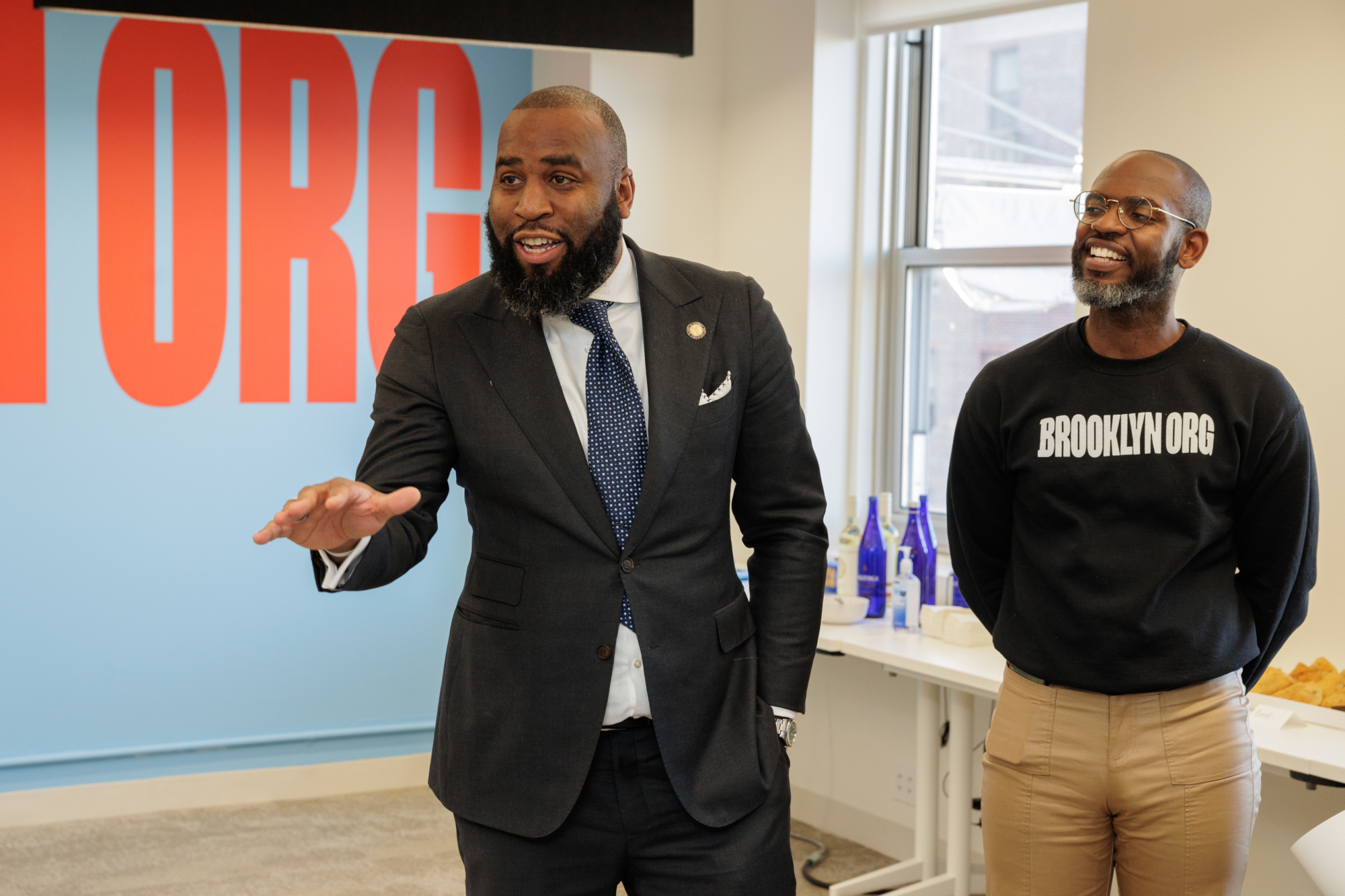 Two men stand in a room, one wearing a suit and the other a sweatshirt with "BROOKLYN.ORG" on it. A large red and blue sign is in the background.