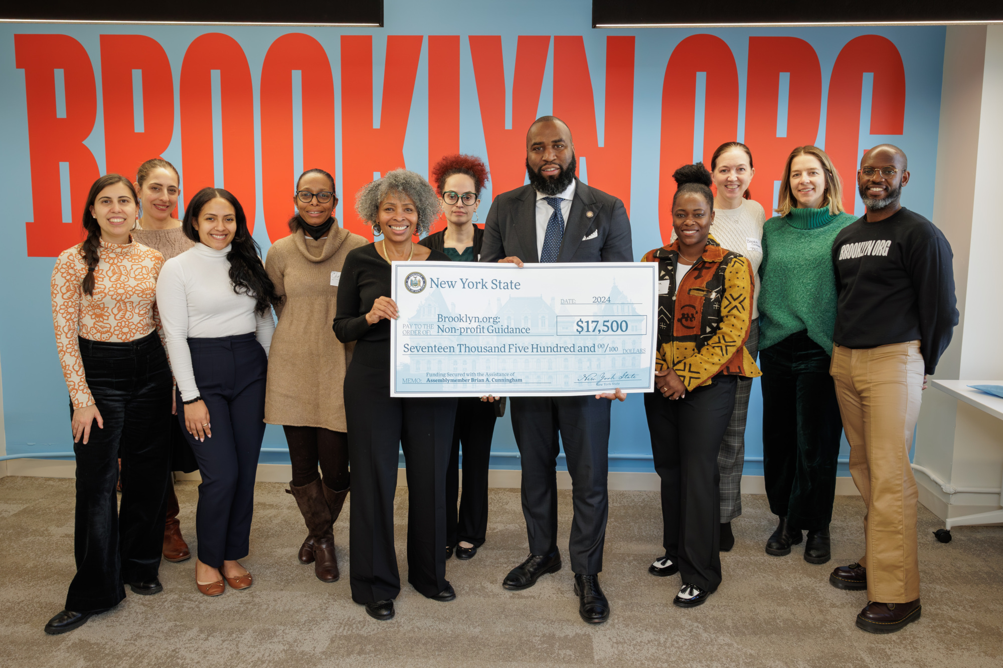 A group of people stands together in an office holding a large check for $17,500, with a red "Brooklyn Org" sign in the background.