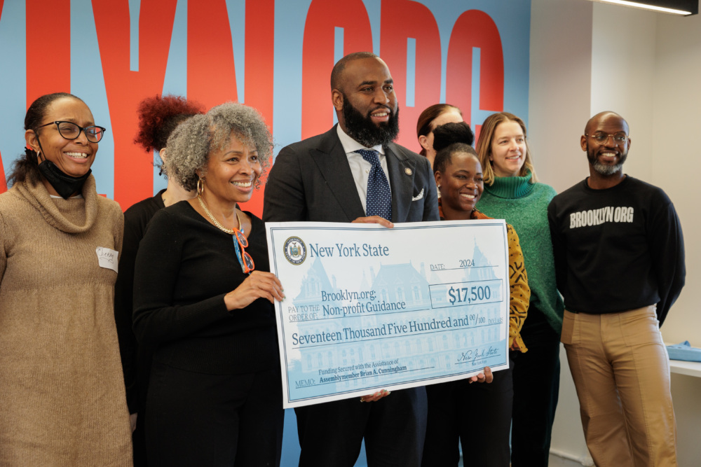 A diverse group of people smiling and holding a large ceremonial check for $17,500 from New York State, with a colorful wall in the background.